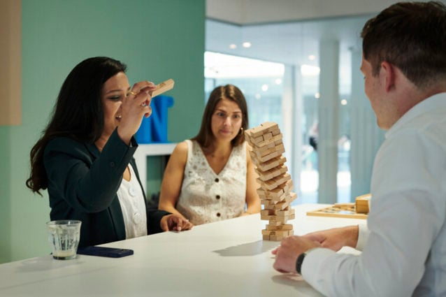 Munter stemning, men med stor spenning, i møterommet når en dresskledd kvinne drar ut en Jenga-brikke som får tårnet til å falle