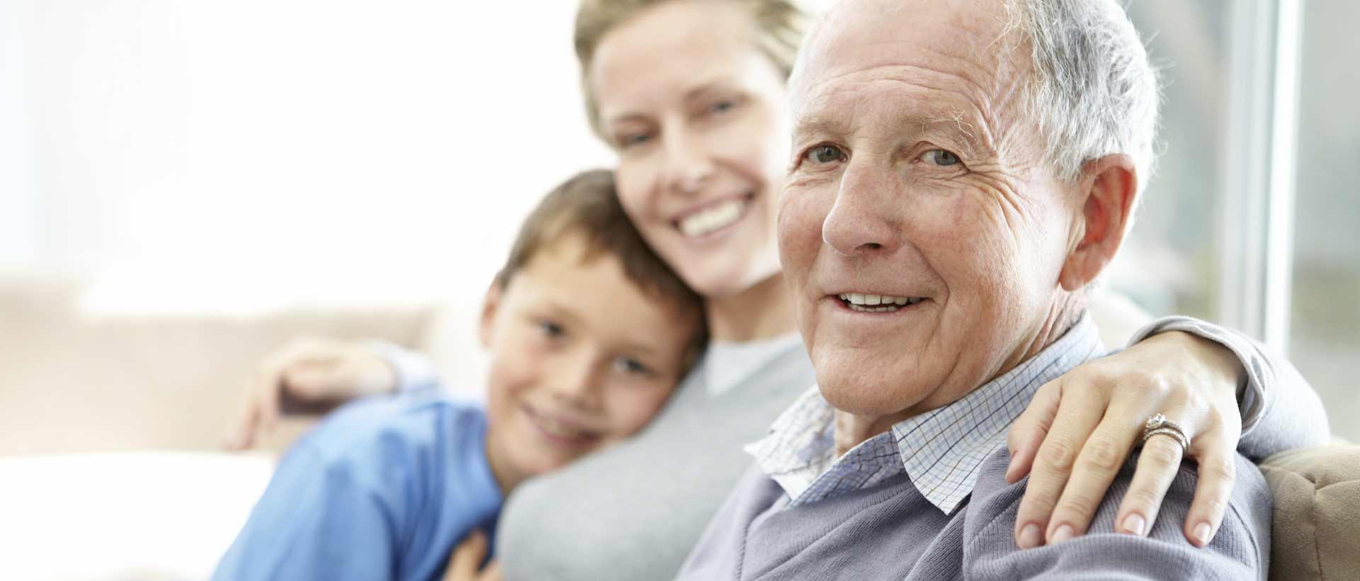 Tre generasjoner smilende sammen på en sofa, med fokus på den eldre mannen, symboliserer hvordan Digihelse forenkler kommunikasjonen mellom eldre, pårørende og helsepersonell gjennom brukervennlig digital teknologi.