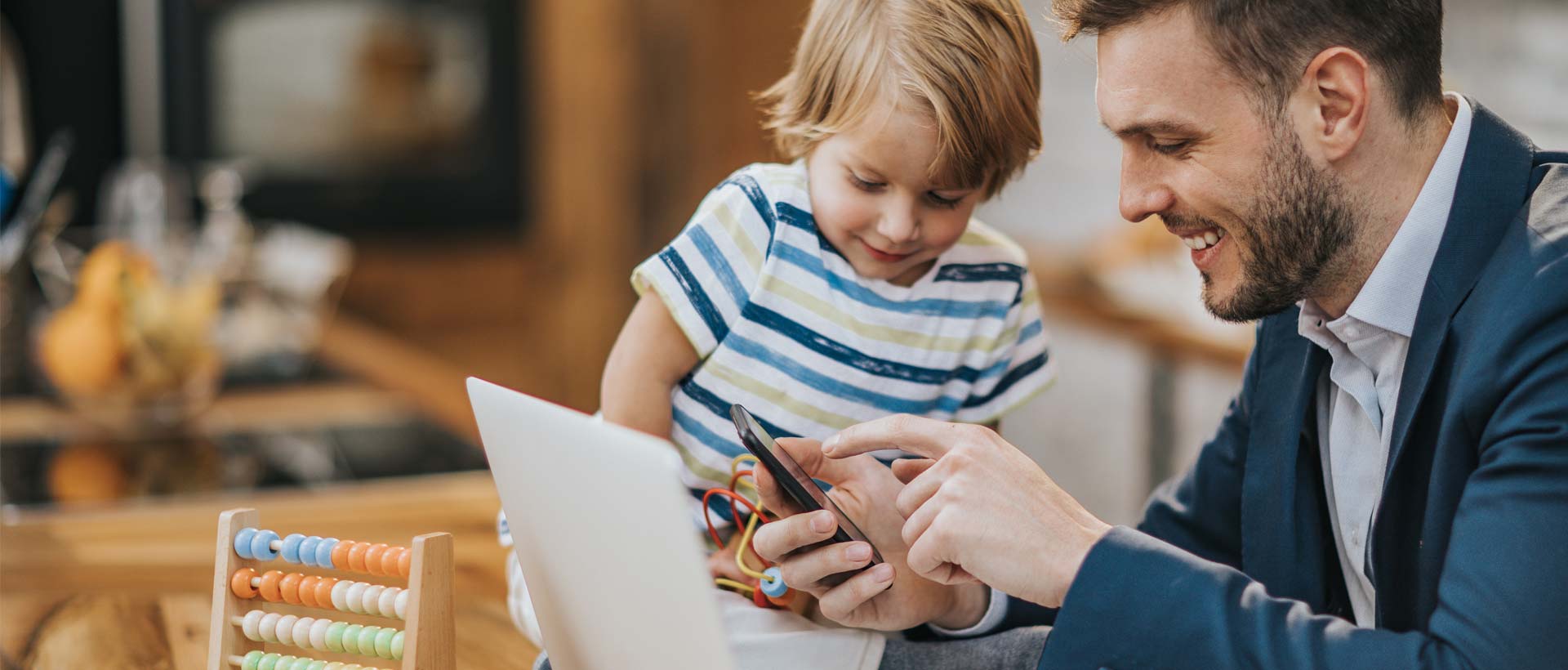 En far bruker Min Barnehage-appen på mobilen sammen med sitt barn ved et bord med et læringsverktøy, og illustrerer hvordan appen forbedrer kommunikasjonen mellom ansatte og foreldre.