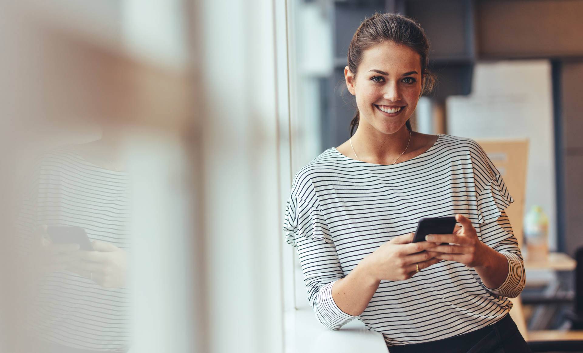 Smilende dame som lener seg inntil en vegg og holder en smarttelefon i hendene