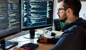 Mannlig utvikler jobber med koding på to skjermer i et moderne kontor, fokusert på programmering relatert til cybersikkerhet.