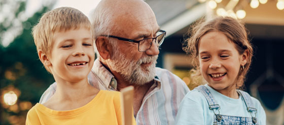 Uformell og koselig samling av en familie i en hage. En eldre mann med briller og skjegg sitter med to barn på fanget hans, begge smiler bredt. I forgrunnen serveres det mat, og i bakgrunnen skimtes et hus med lyslenker. Bildet illustrerer varme og samvær, reflekterer Visma Flyt sine fagløsninger med fokus på mennesker.