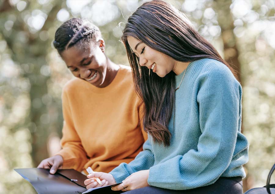 To unge kvinner sitter utendørs og smiler mens de samarbeider med dokumenter i en mappe til voksenopplæring. Den ene har på seg en oransje genser og den andre en blå, og de ser avslappet og engasjert ut i samtale.